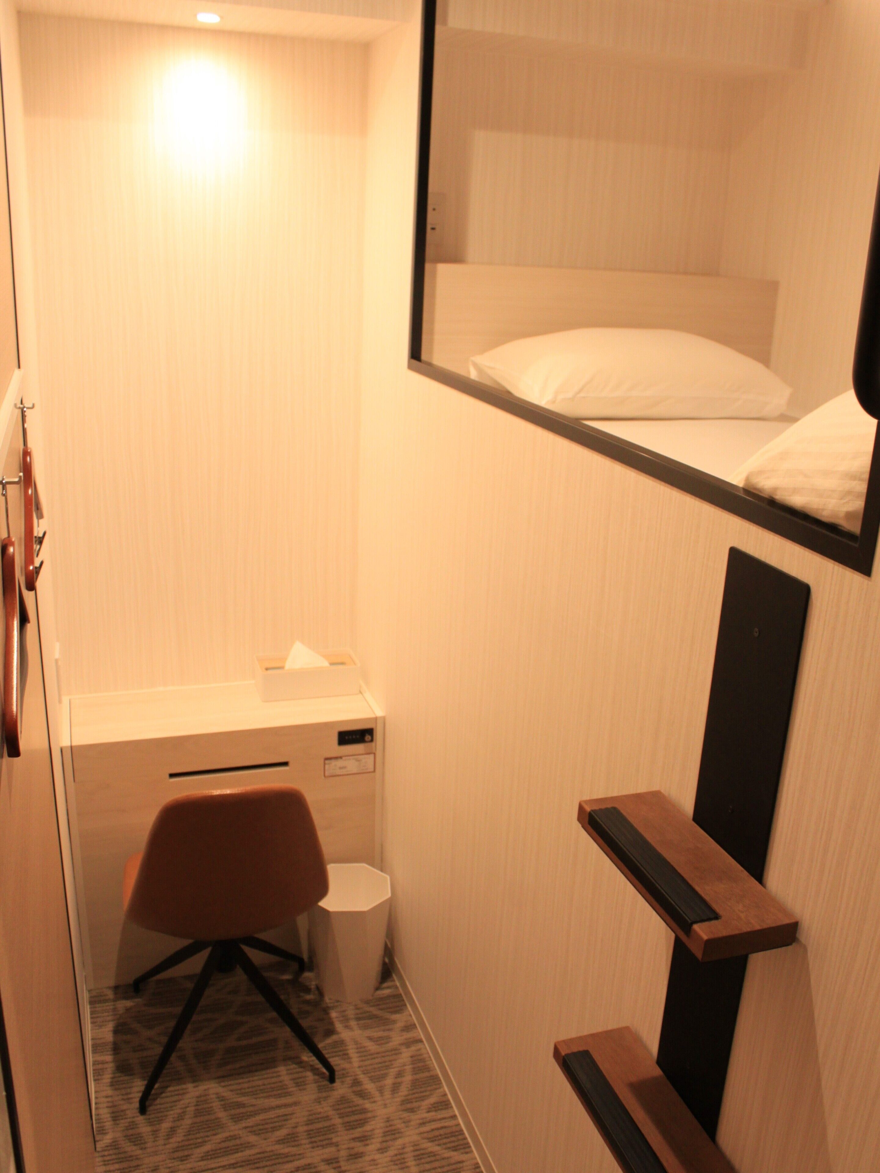 A compact hotel room with a bunk bed, a desk with a chair, a small trash bin, and a mirror.