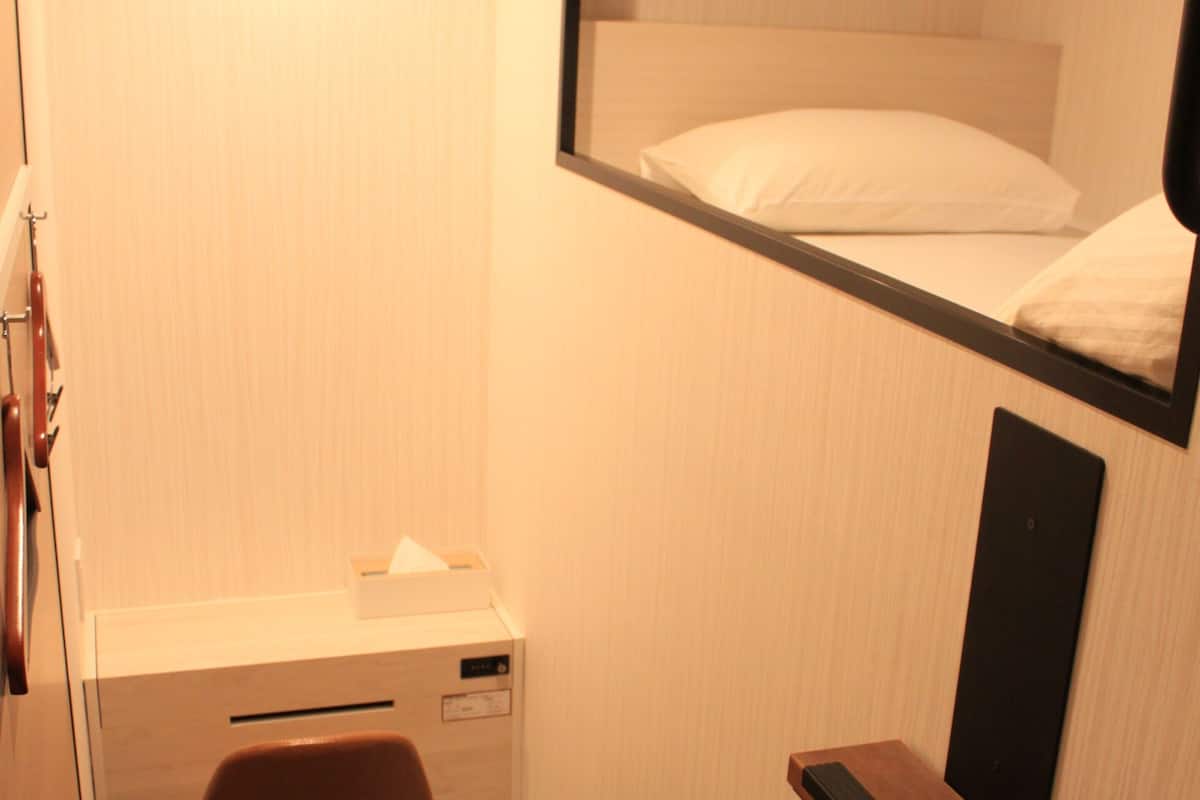 A compact hotel room with a bunk bed, a desk with a chair, a small trash bin, and a mirror.