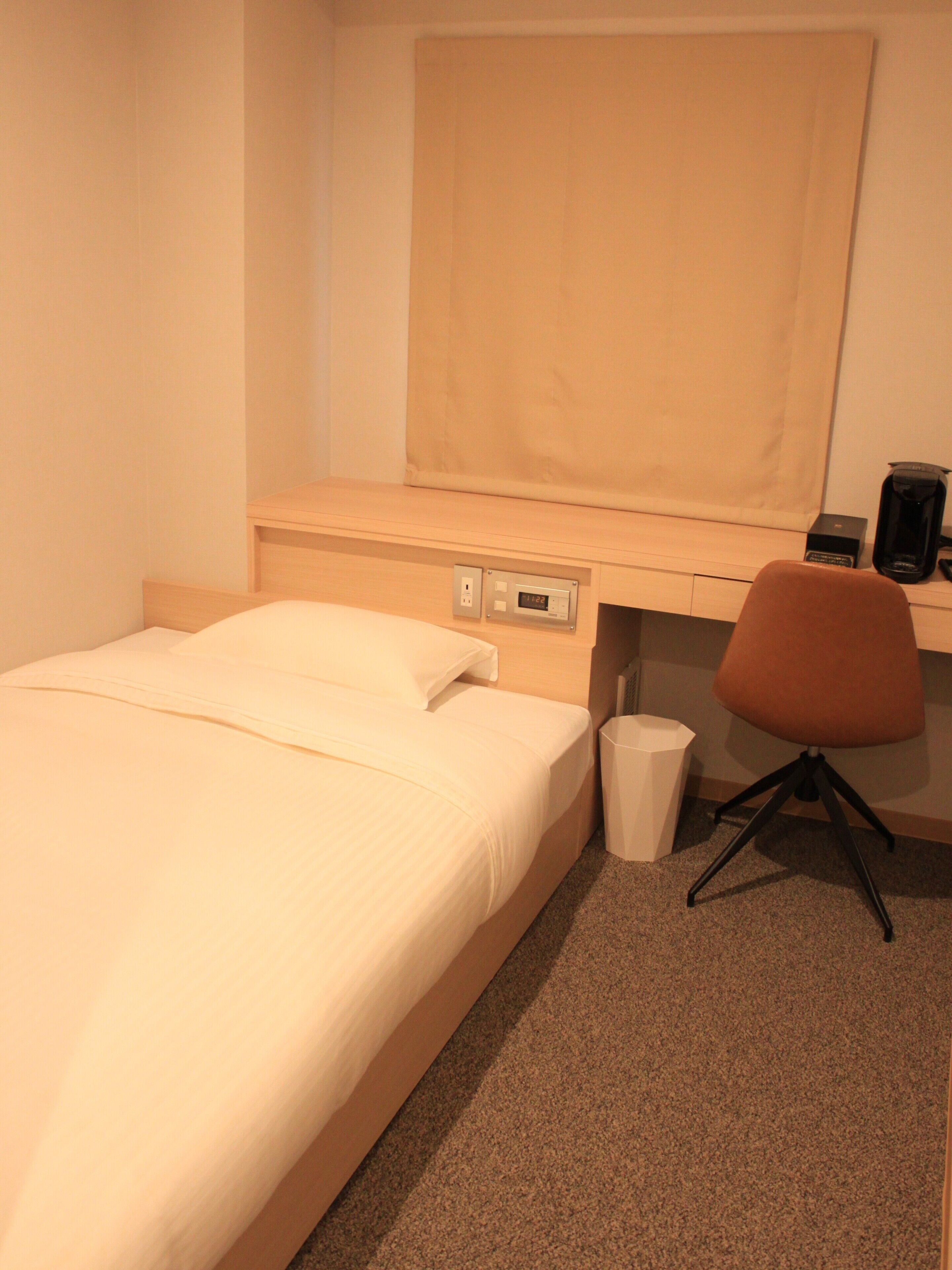 A hotel room with a bed, a desk, a chair, a trash bin, and a coffee maker.