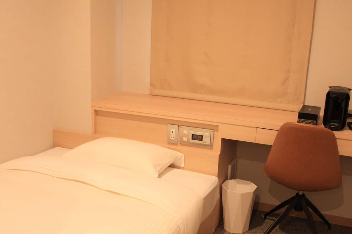 Una habitación de hotel con cama, escritorio, silla, cesto de basura y cafetera.