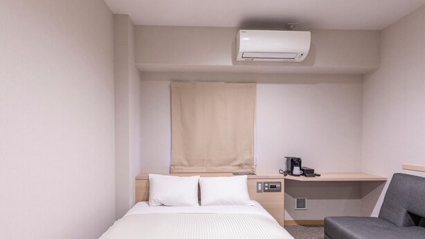 Habitación de hotel pequeña con cama, escritorio, silla, cafetera y aire acondicionado.
