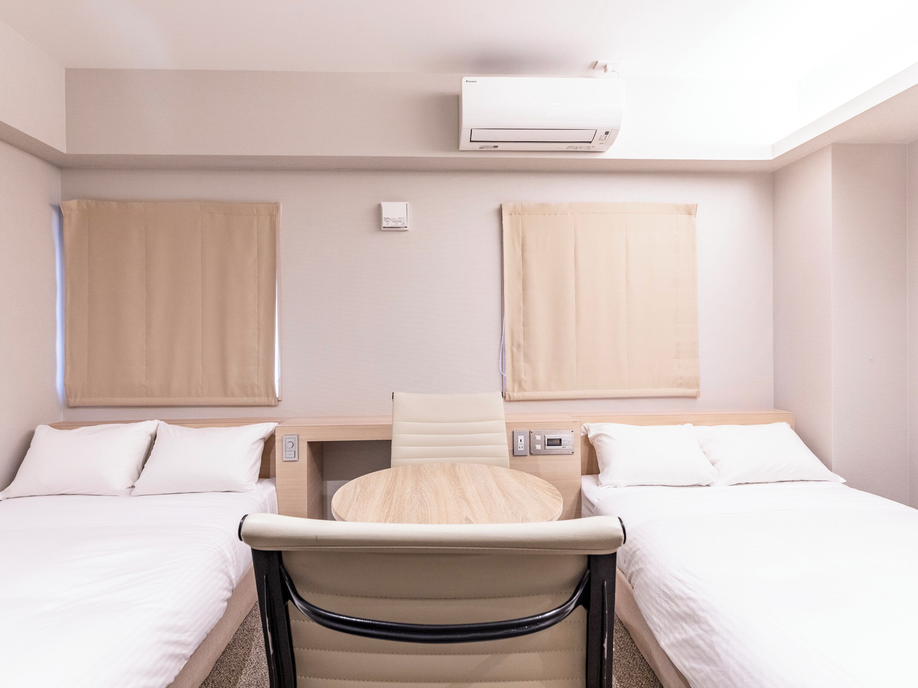 Une chambre d’hôtel avec deux lits, une table ronde, une chaise, un appareil de climatisation et une fenêtre avec des rideaux.