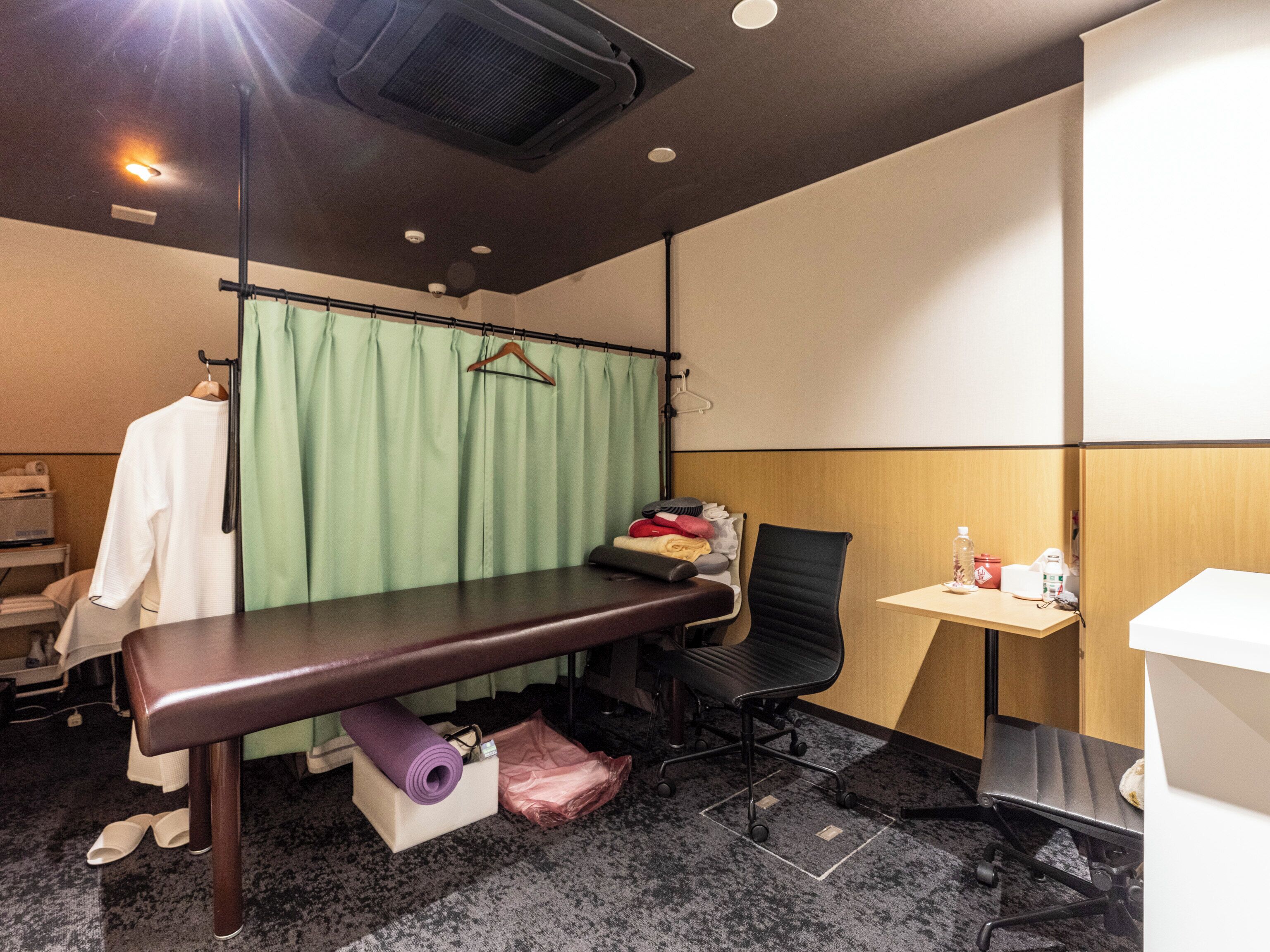 A room with a massage table, a chair, a small table with a bottle, and a green curtain.
