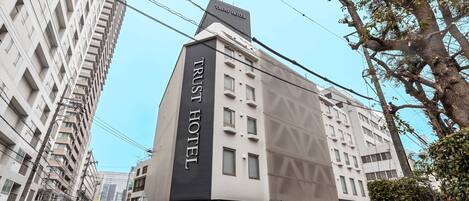 Un hotel moderno con un letrero destacado que dice 'TRUST HOTEL', varios pisos y un estacionamiento con coches y motocicletas.