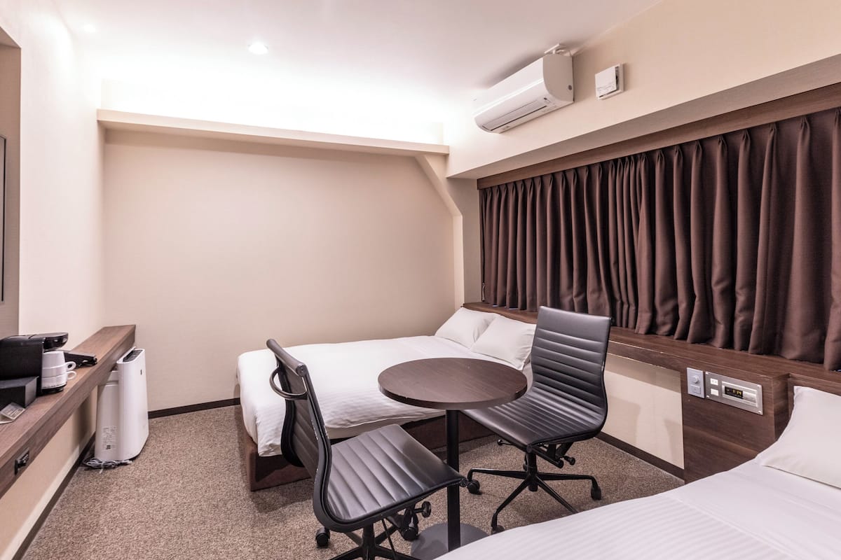 Habitación de hotel compacta con una cama, un escritorio con silla, una mesita y un equipo de aire acondicionado en la pared.
