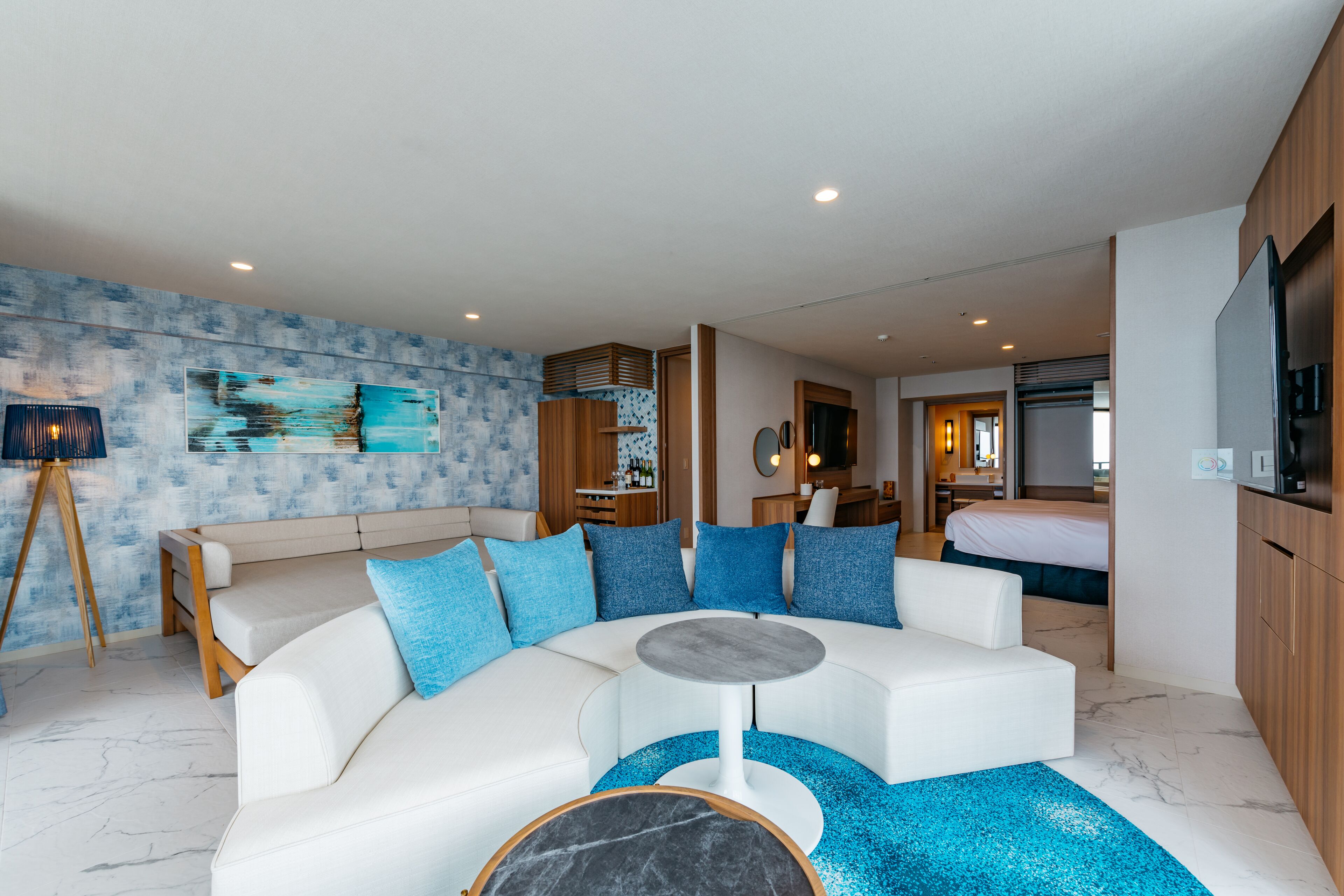 A modern living room with a white sectional sofa, a round coffee table, and a blue accent rug. There is a kitchen area visible in the background with a refrigerator and a microwave.