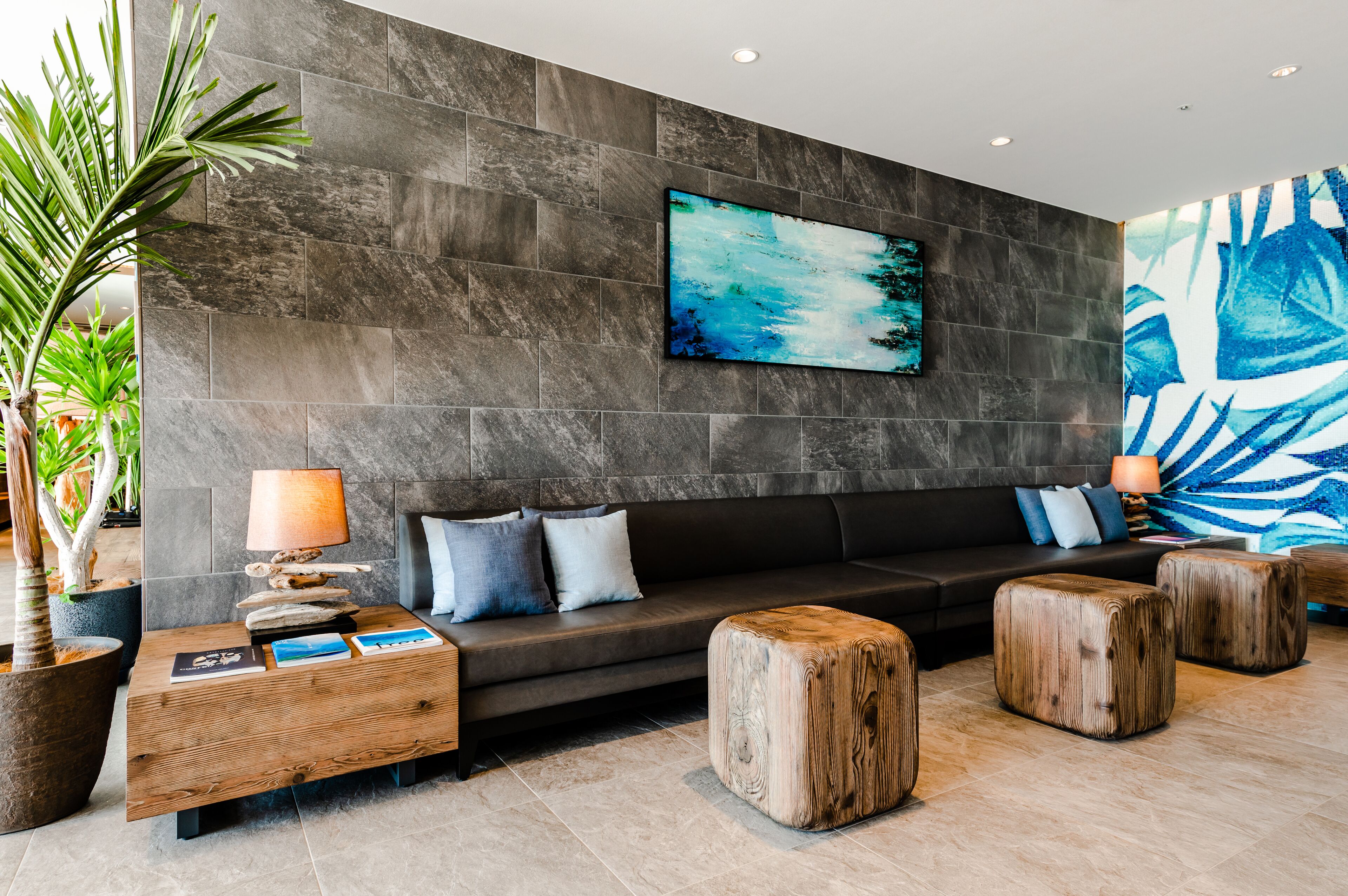 A modern lobby with a dark stone wall, a black sofa with cushions, wooden side tables, and a large abstract painting.