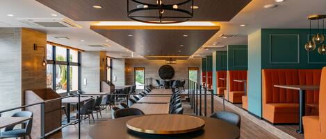 A modern indoor space with a long dining table, multiple seating areas, and a central circular light fixture.