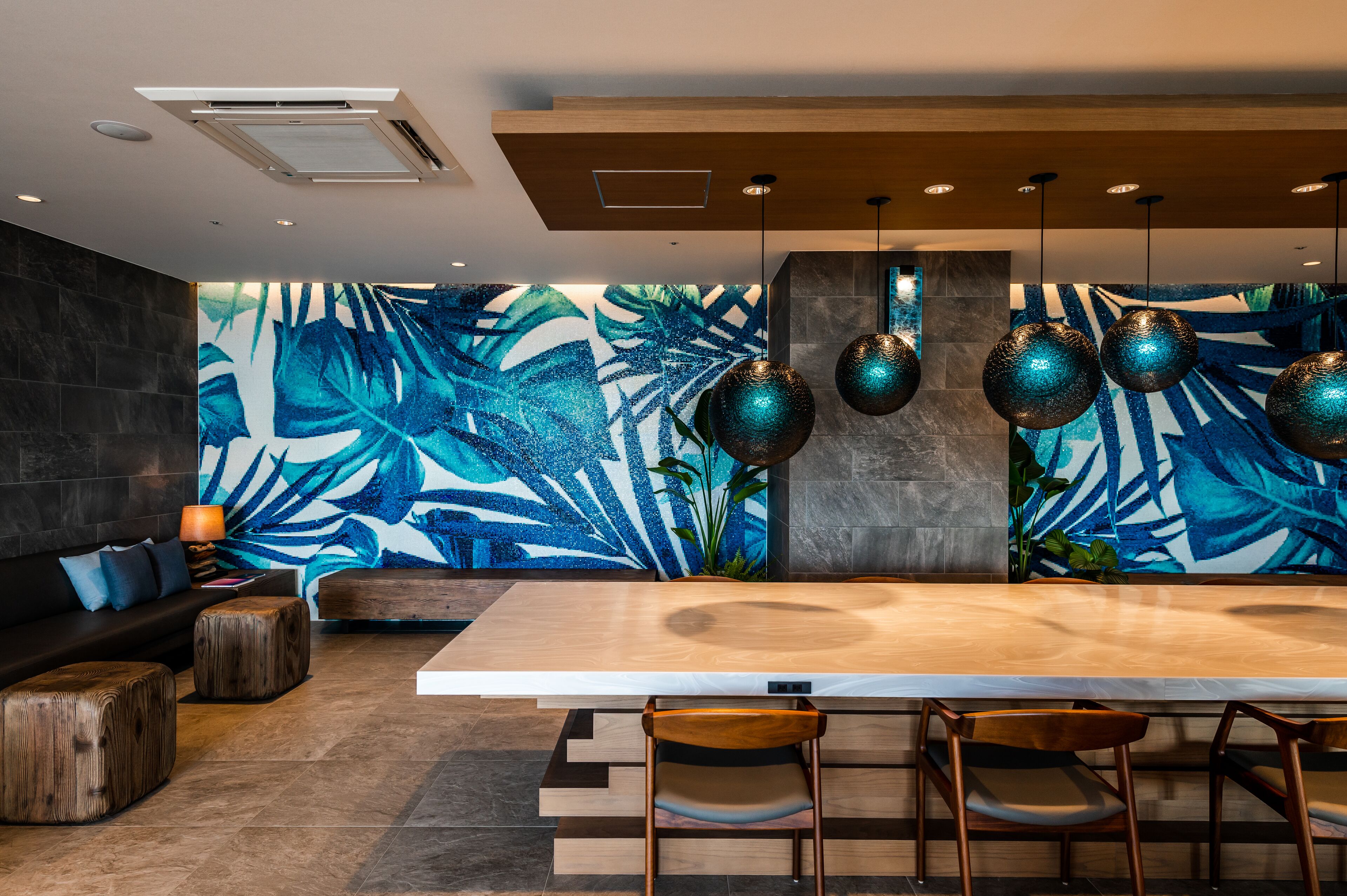 A modern dining area with a large wooden table, three chairs, a dark sofa, and a wall mural with blue and green tropical leaves.