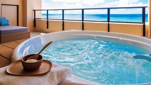 Un bain à remous avec une cuillère en bois et un bol posés sur un plateau, un fauteuil confortable et un balcon offrant une vue sur l’océan.