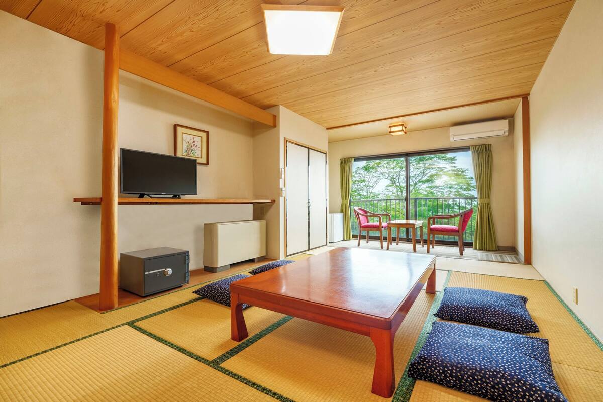 Ein traditioneller japanischer Raum mit Tatami-Böden, einem niedrigen Holztisch, einem Fernseher und einer Schiebetür zu einem Balkon mit Blick auf Grünflächen.