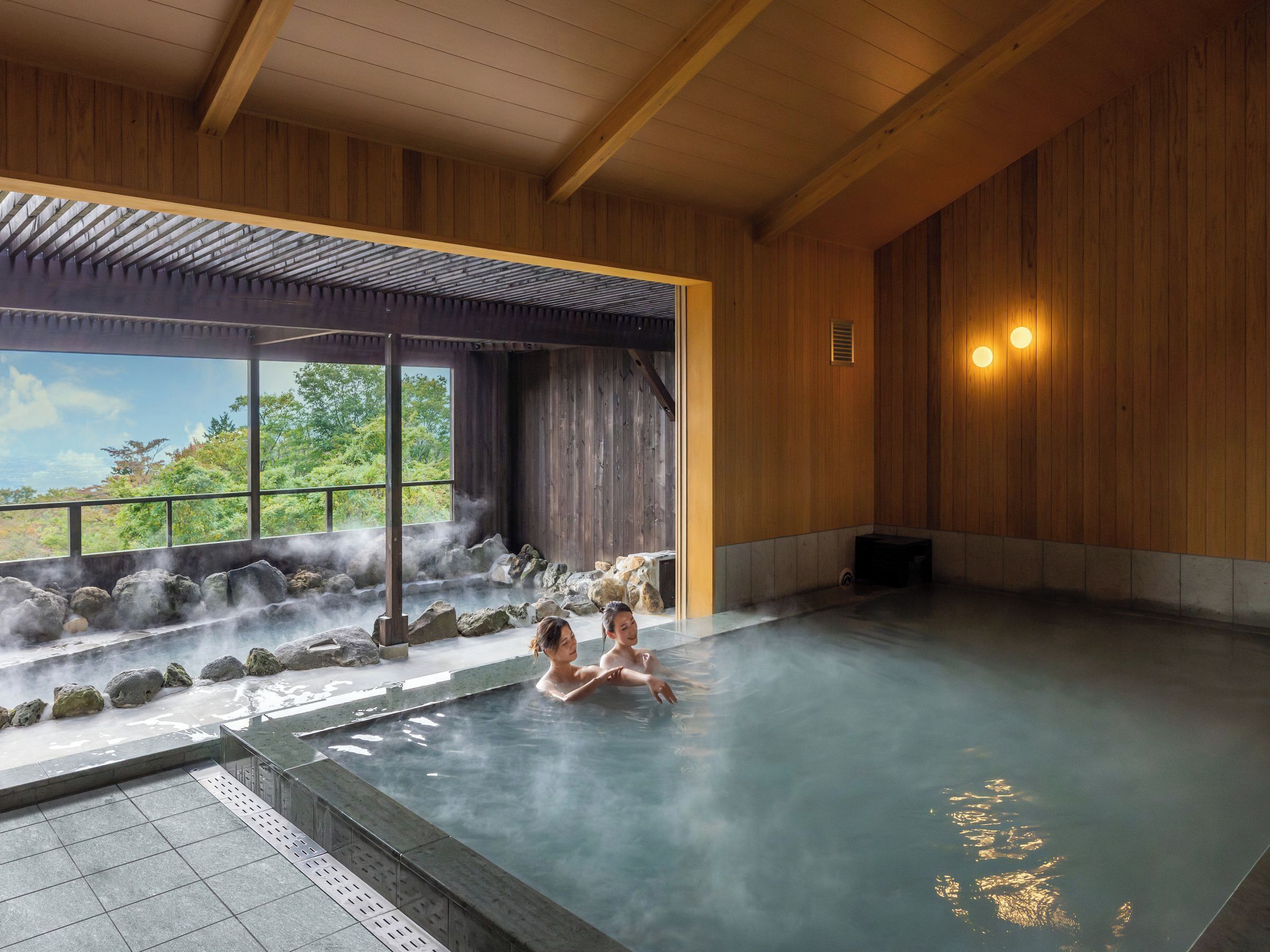 A traditional Japanese onsen with a view of the outdoors, a stone bath area, and wooden interiors.
