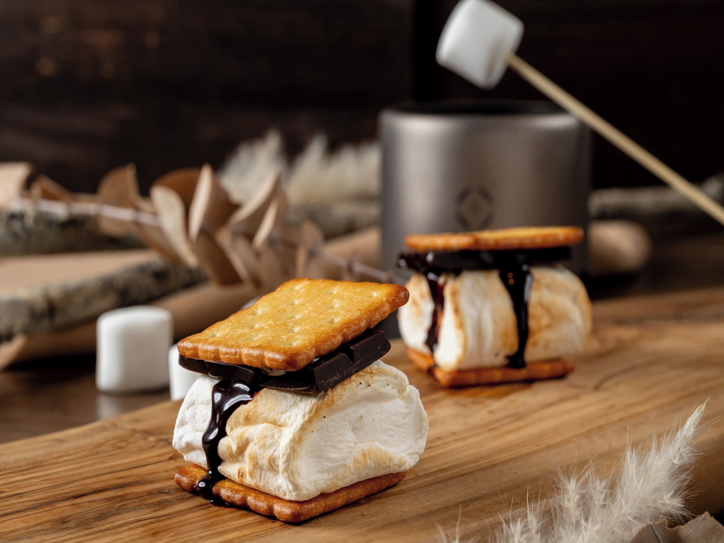 Deux sandwichs à la crème glacée, nappés de sauce au chocolat et garnis de guimauves, posés sur une surface en bois.