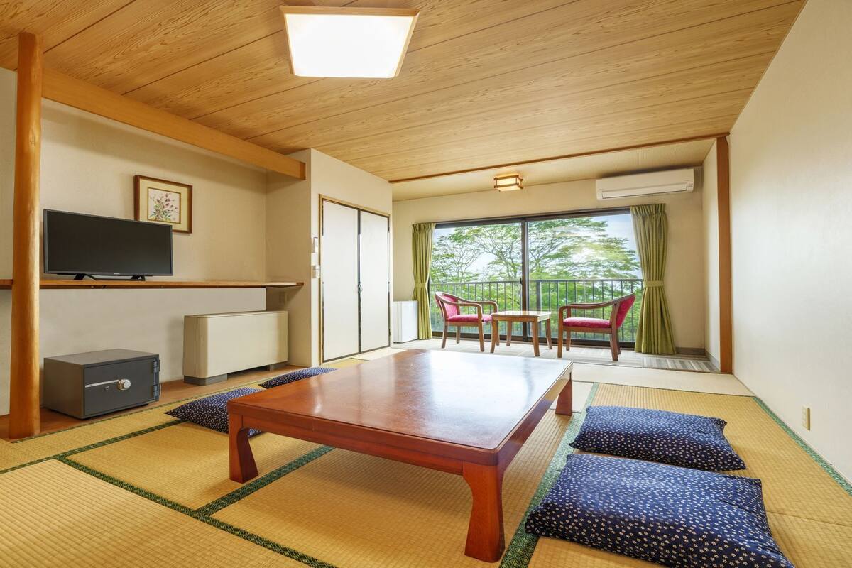Ein traditioneller japanischer Raum mit Tatami-Böden, einem niedrigen Tisch, einem Fernseher und einer Schiebetür zu einem Balkon mit Aussicht.