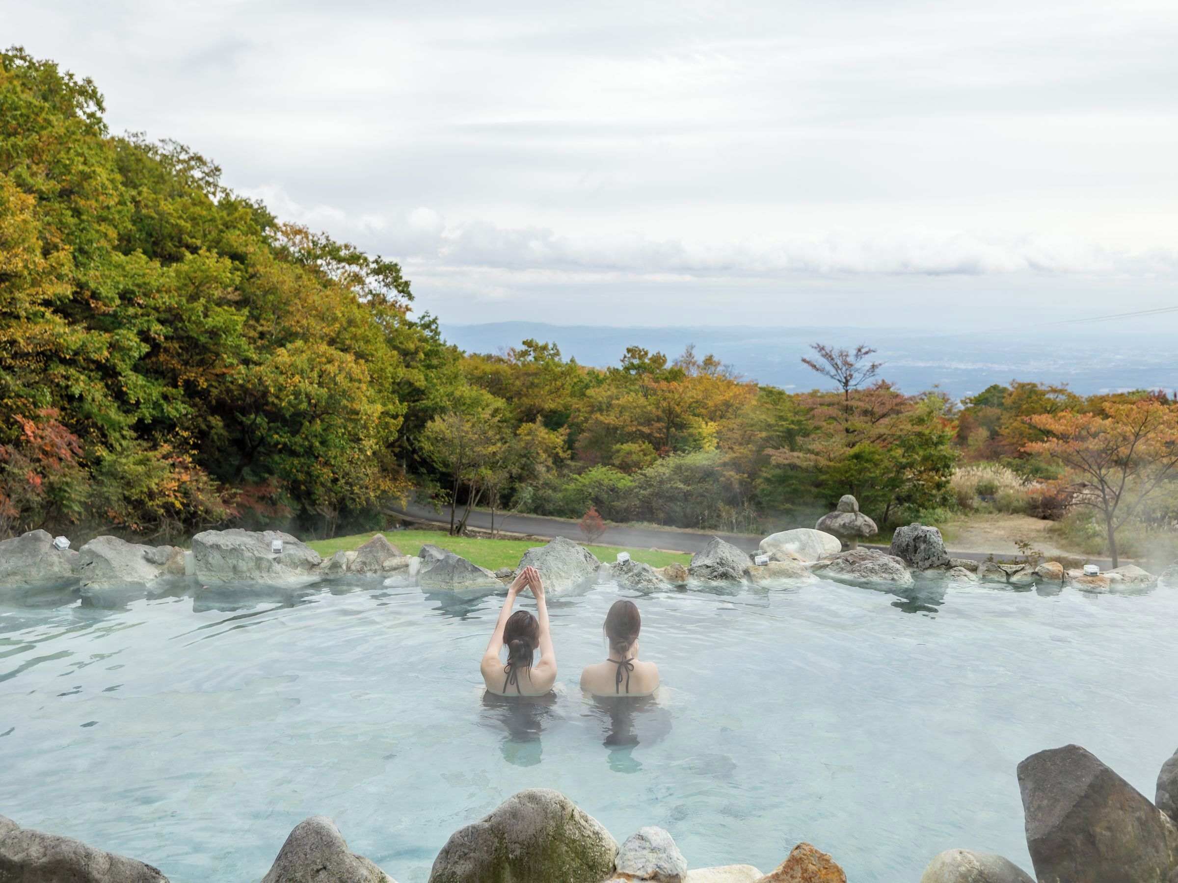 Two people in a clear, rocky hot spring surrounded by trees and a road in the background.