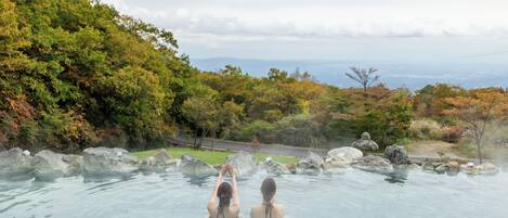Two people in a clear, rocky hot spring surrounded by trees and a road in the background.