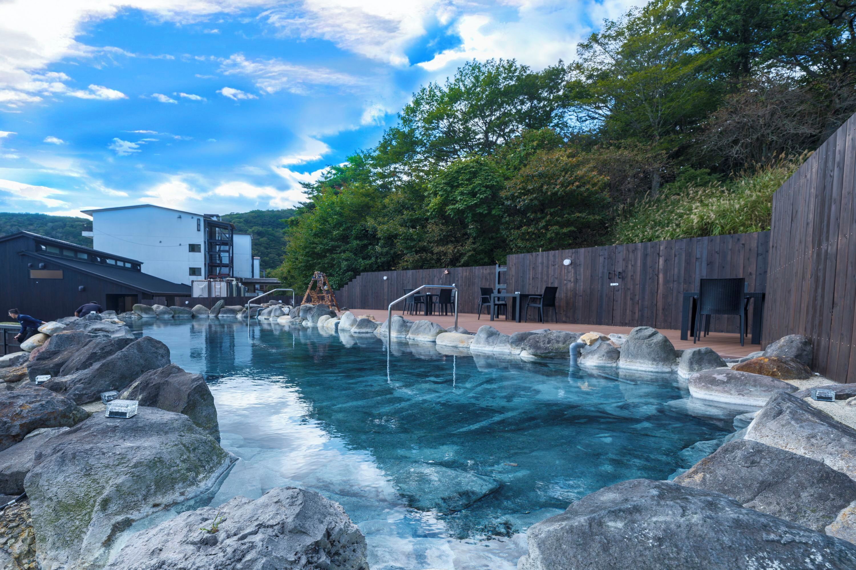 Una piscina cristalina con rocas, terraza de madera y mobiliario exterior.