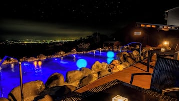 A nighttime outdoor pool area with a view of the city skyline, a wooden deck, and a lounge chair.
