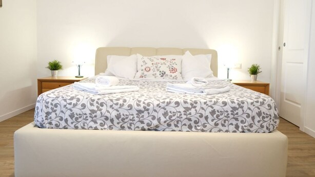Een net opgemaakt bed met een bloemenprint deken en kussens, twee nachtkastjes met lampen en kleine potplanten, en een houten vloer.