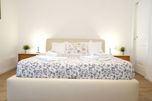 Een net opgemaakt bed met een bloemenprint deken en kussens, twee nachtkastjes met lampen en kleine potplanten, en een houten vloer.