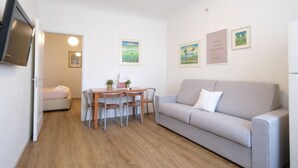 Een compacte, moderne kamer met een bank, een eettafel met stoelen, een flatscreen-televisie aan de muur en een bed op de achtergrond.
