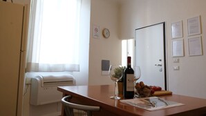 Een kleine, goed verlichte kamer met een kitchenette, een eettafel met een fles wijn en glazen, en een raam met gordijnen.