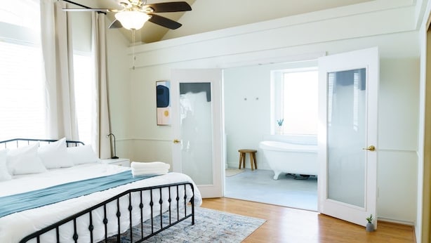 Una camera da letto con un letto, una ventola a soffitto, una finestra e un bagno visibile attraverso una porta aperta.
