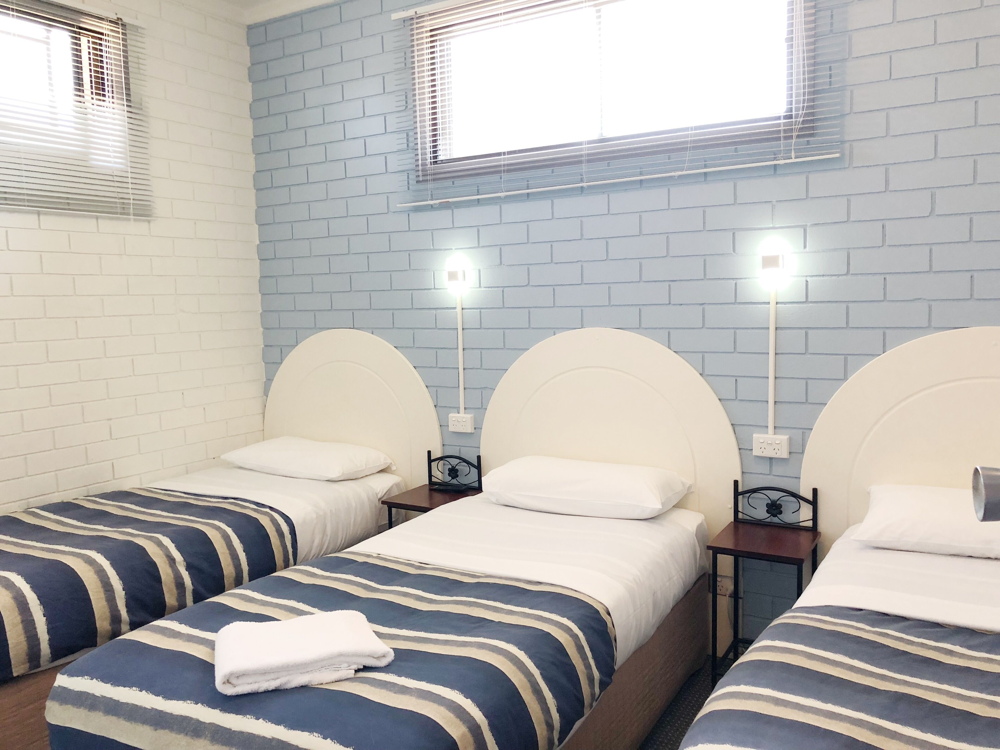 A hotel room with two single beds, each with a striped bedspread and white pillows. There are two bedside tables with lamps, and the walls are covered with blue brick-pattern wallpaper.
