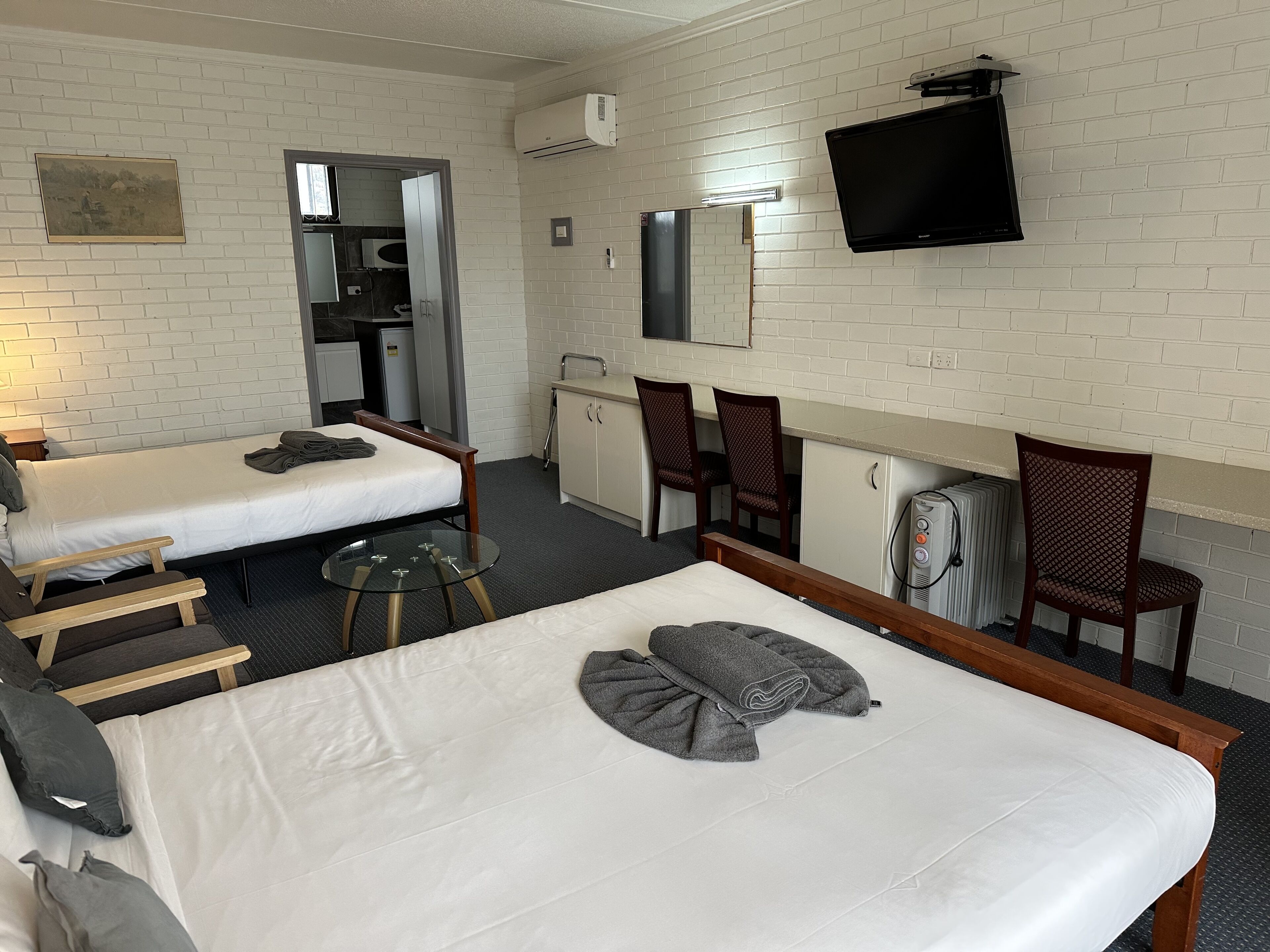 Una habitación de hotel con dos camas individuales, una kitchenette pequeña, un televisor montado en la pared y un espejo sobre el lavamanos.