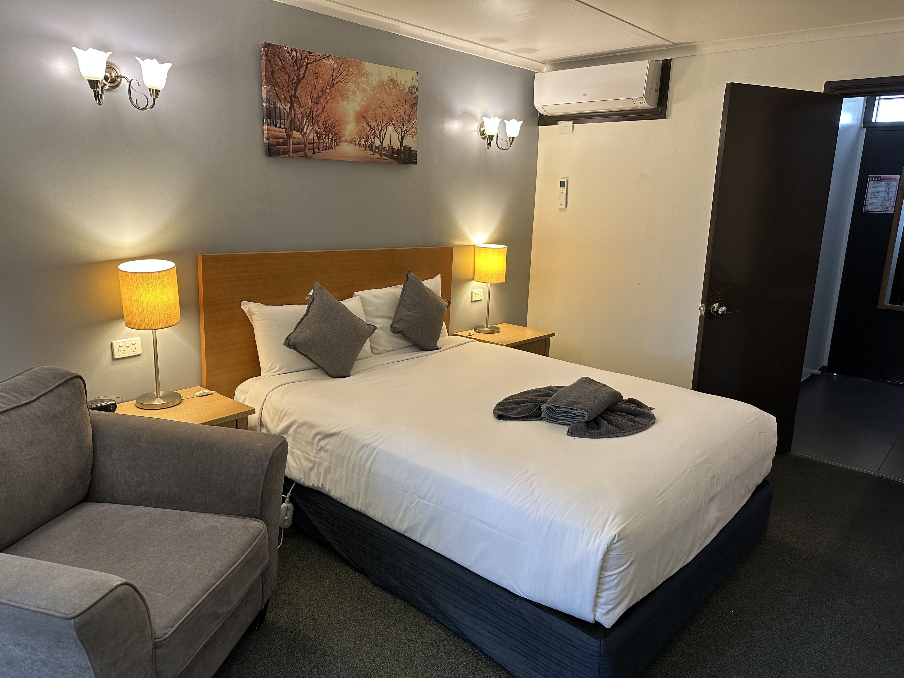 Una habitación de hotel con cama, dos lámparas de noche, aire acondicionado en la pared y un cuadro de una calle arbolada.