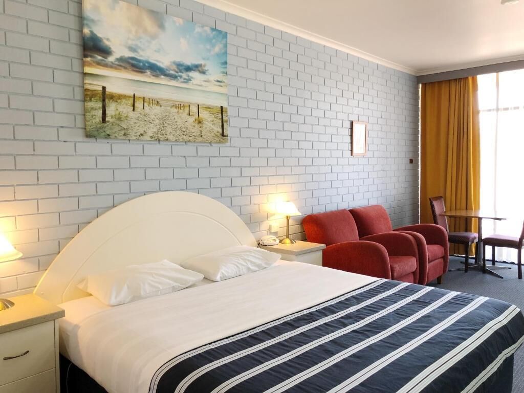 A hotel room with a white brick wall, a large bed with a striped bedspread, a painting of a beach scene, red chairs, and a table with yellow curtains.
