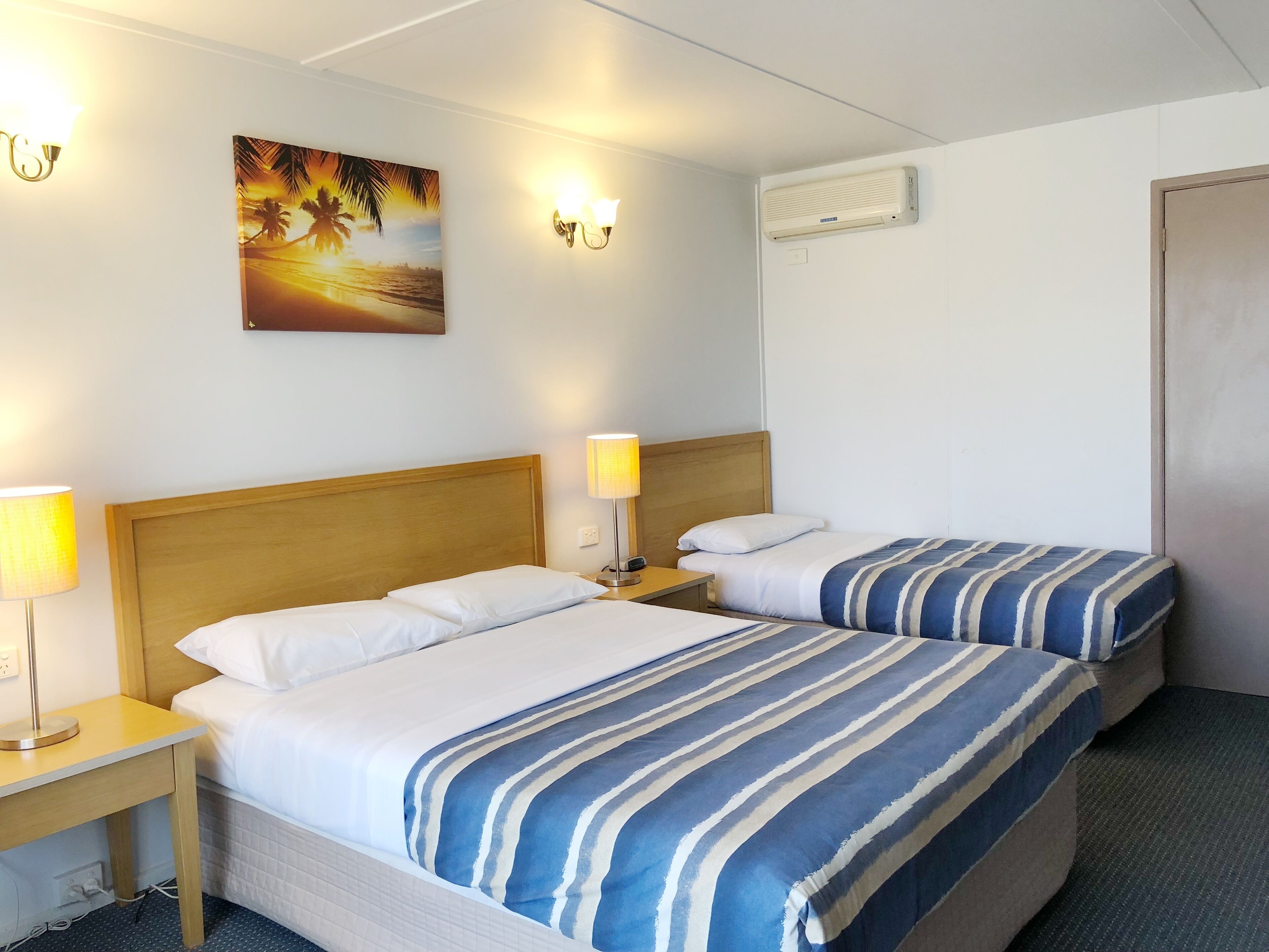 A hotel room with two beds, a nightstand with a lamp, a wall-mounted air conditioner, and a sunset painting on the wall.