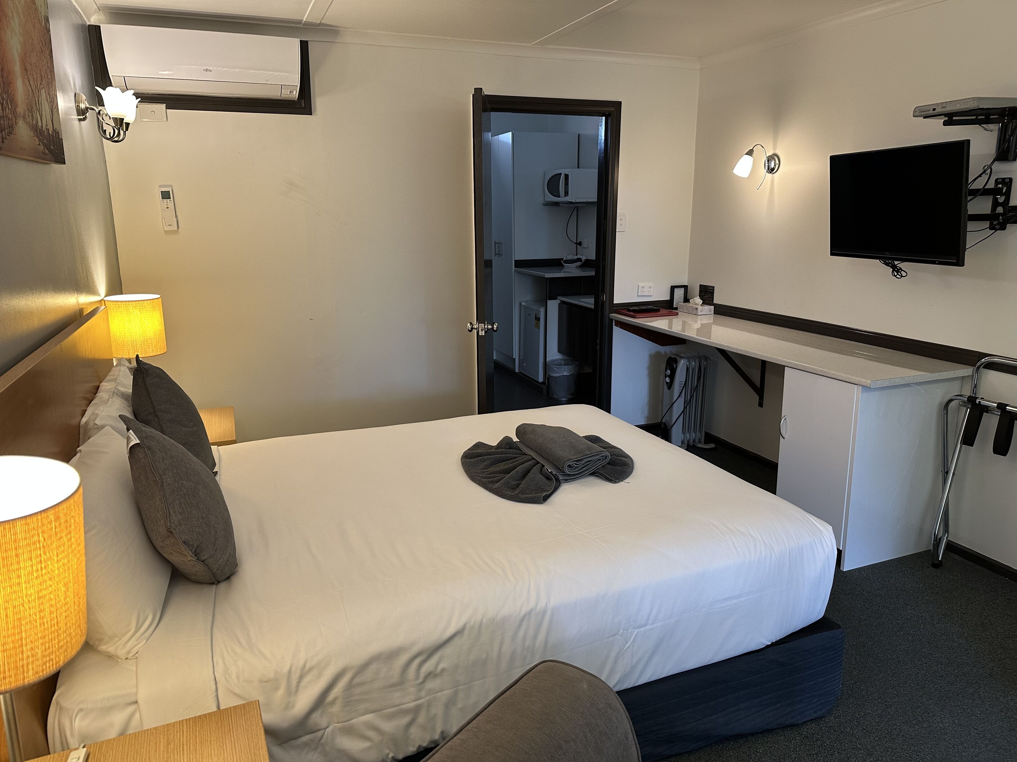 A hotel room with a bed, bedside lamps, a flat-screen TV mounted on the wall, and a small kitchenette area.