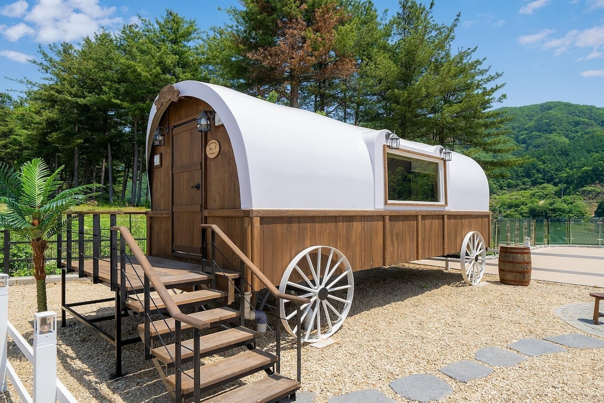 Une cabane de style couvert, avec une façade en bois et un toit blanc, située sur un chemin de gravier. Un escalier en bois mène à l’entrée.