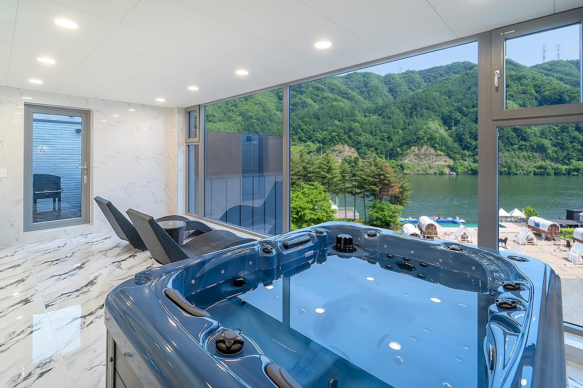 Une salle de spa moderne dotée d’un grand jacuzzi, d’une porte vitrée donnant sur un balcon avec vue sur le lac, et d’un coin salon avec un fauteuil et une petite table.