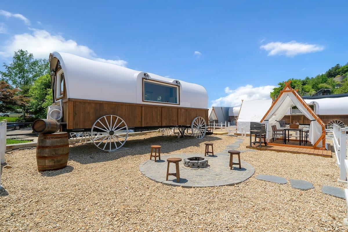 Un chariot couvert et une structure de type tipi se trouvent dans un espace recouvert de gravier, avec des tables et des chaises.