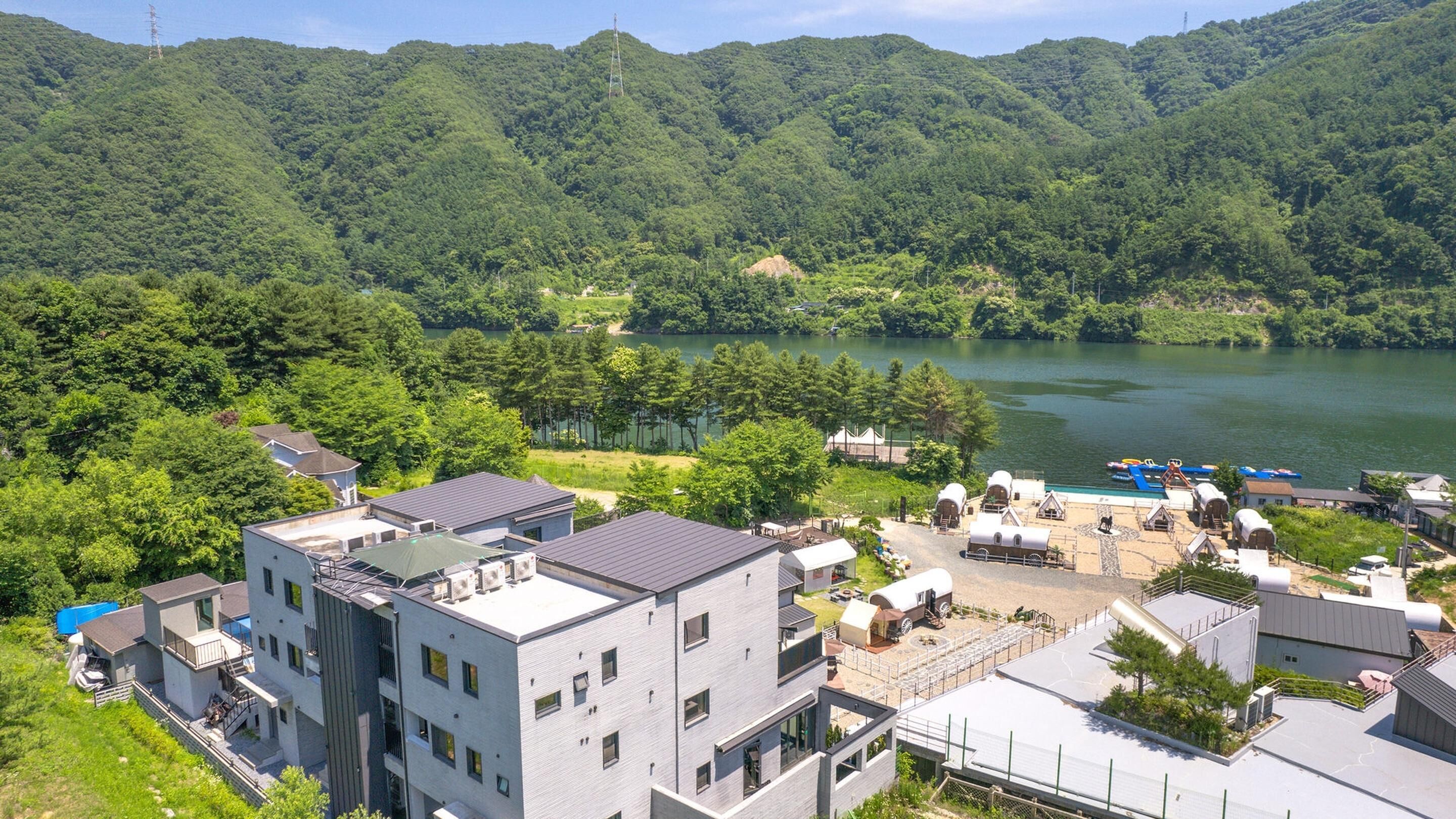 A modern building with multiple floors, surrounded by lush greenery and a lake.