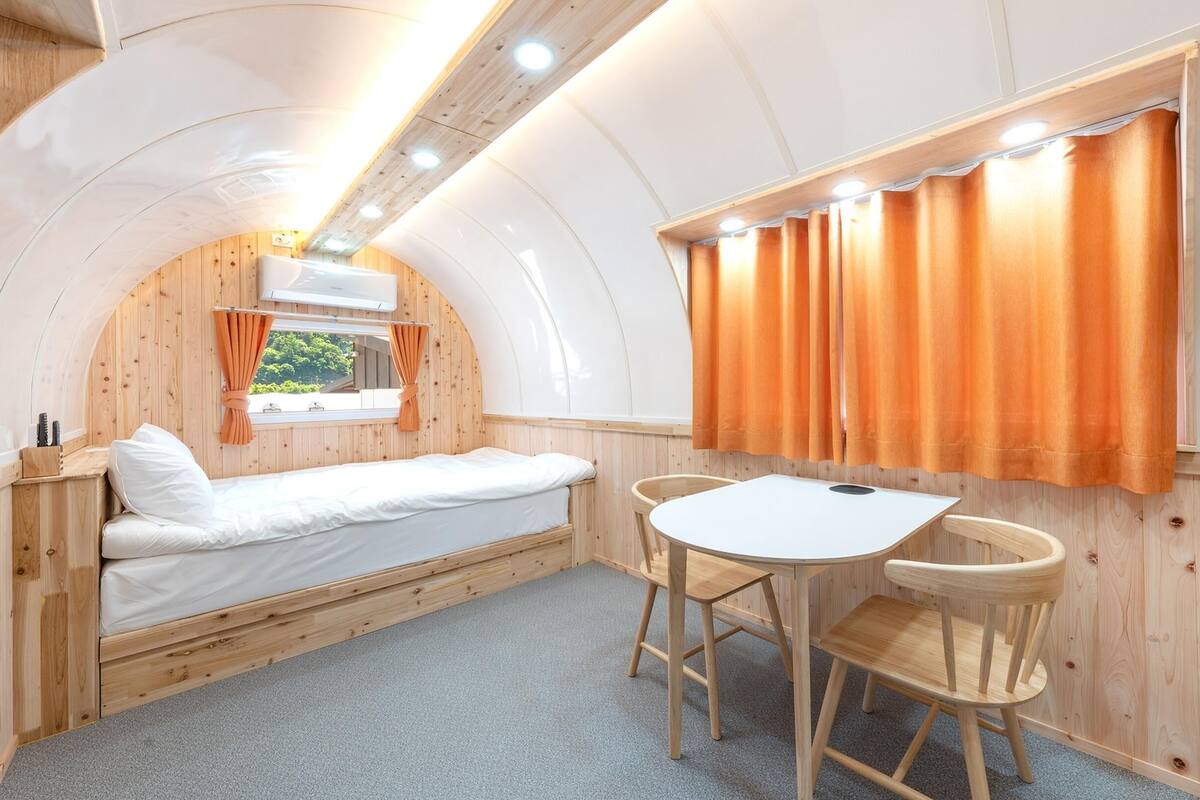 Une petite pièce aux murs lambrés, avec un lit superposé, une petite table et une fenêtre avec des rideaux.