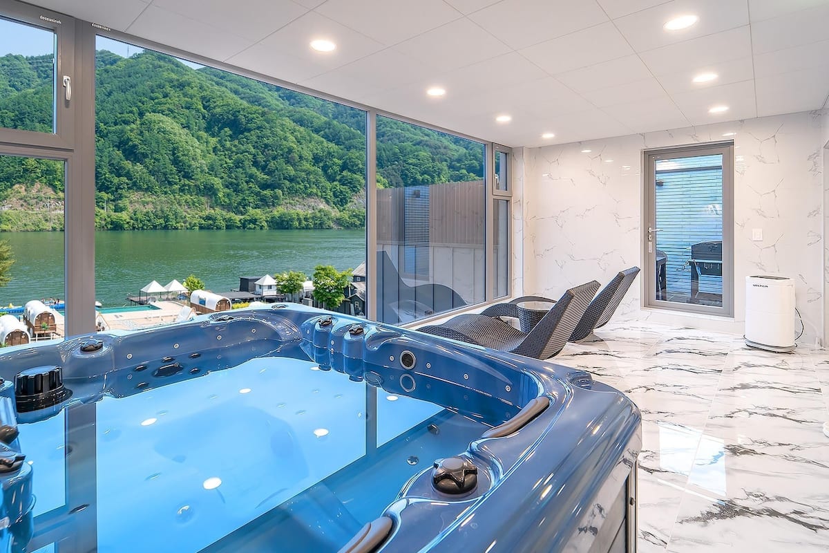 Un espace spa moderne doté d’un bain à remous, de grandes fenêtres et offrant une vue sur un lac et une forêt.