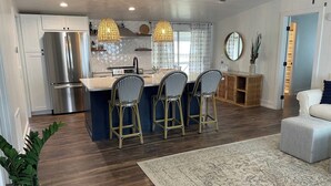 Una cocina moderna con una isla central, taburetes de bar y un comedor con sofá y reposapiés blancos.