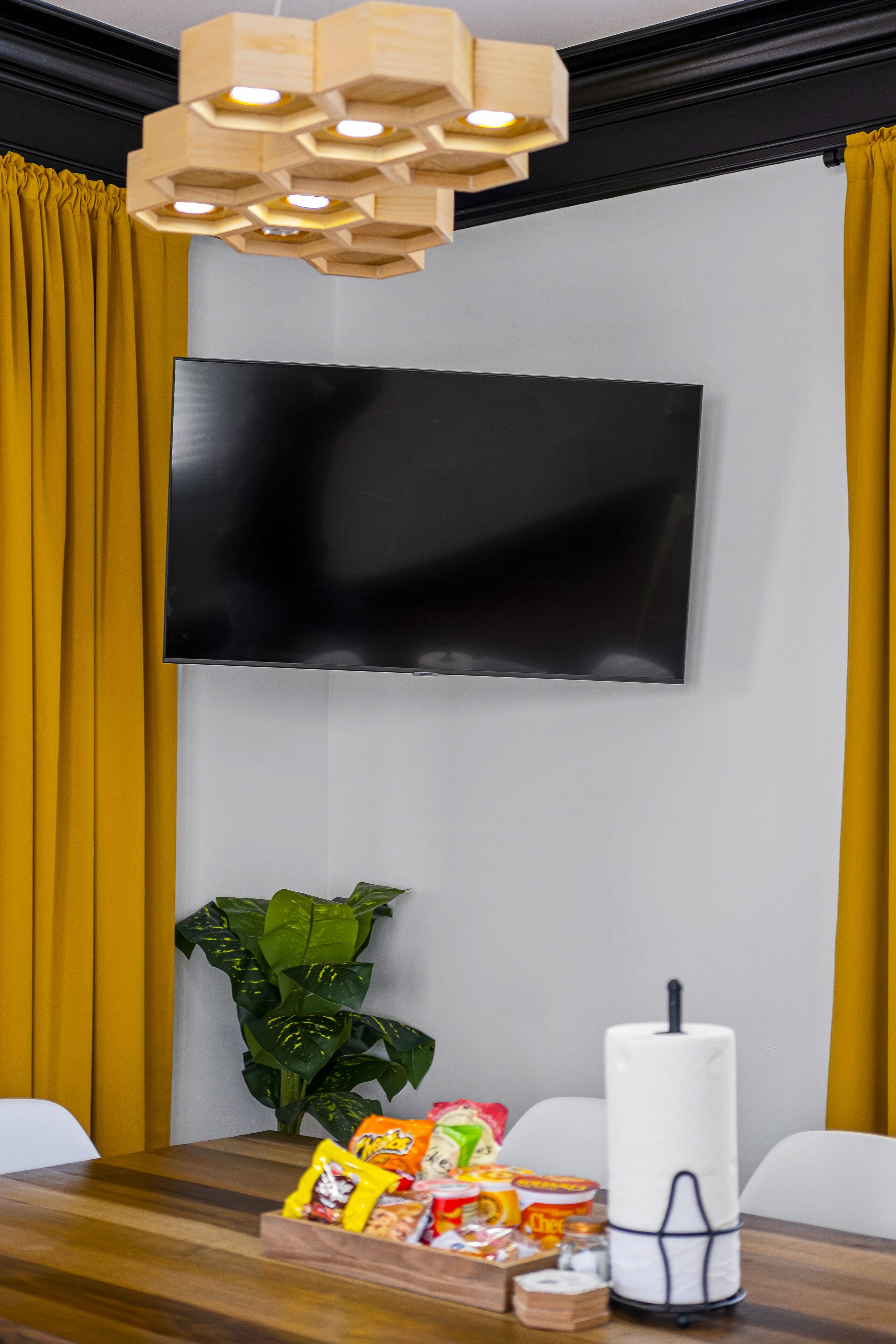 A modern living room with a flat-screen TV mounted on the wall, a wooden table with snacks, a potted plant, and a candle.