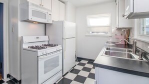 A kitchen with white cabinets, a gas stove, a microwave, a refrigerator, a black and white checkered floor, and a window with blinds. - Shamrock Sanctuary walkable to Notre Dame 1.5 mi (South Bend)