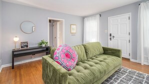 A living room with a green sofa, a round mirror, a console table with a lamp, and a door with a white curtain. - Shamrock Sanctuary walkable to Notre Dame 1.5 mi (South Bend)