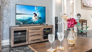 Ett modernt vardagsrum med en stor platt-TV som visar en surfare på en våg, en träinredd TV-möbel med ett glasskåp, två vinglas på en underlägg och en vas med blommor.