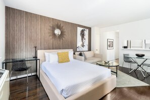 A modern hotel room with a large bed, a glass desk, a sofa, and a dining area.