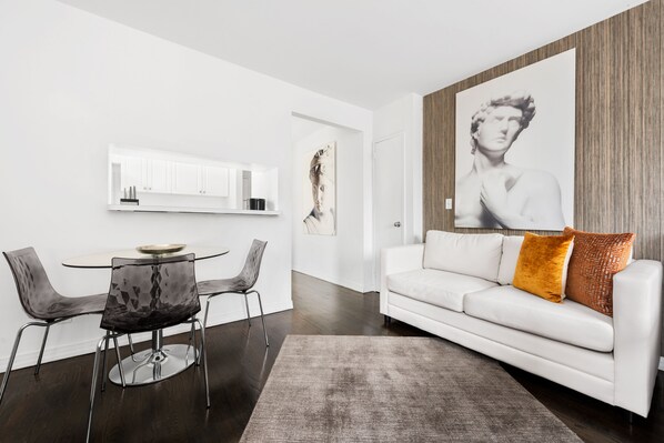 A modern living room with a white sofa, a glass coffee table, and a dining area with a white cabinet and chairs.