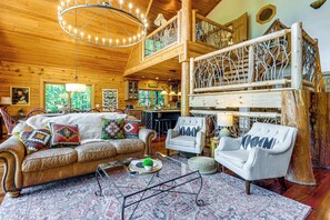 A log cabin interior with a high ceiling, a wooden staircase, a sofa, a chair, and a dining area. - Smoky Mtn Cabin by Cataloochee Valley Elk Viewing! (Waynesville)