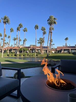 Een vuurplaats in een ronde plantenbak, een zwembad en uitzicht op een resort met palmbomen en gebouwen.