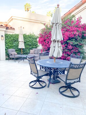 An outdoor patio with a round table and chairs, surrounded by lush greenery and vibrant pink flowers. - Palm Valley Getaway with all the amenities & golf water view! (Palm Desert)