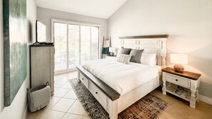 A bedroom with a large bed, a bedside table, a nightstand, a lamp, a window with blinds, and a view of the outdoors. - Palm Valley Getaway with all the amenities & golf water view! (Palm Desert)
